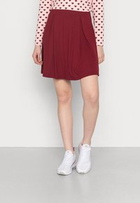 Bordeauxrode skaterrok met plooien aan de taille, gemaakt van zachte stof. Gecombineerd met witte sportschoenen en een roze top met lange mouwen en hartjespatroon.