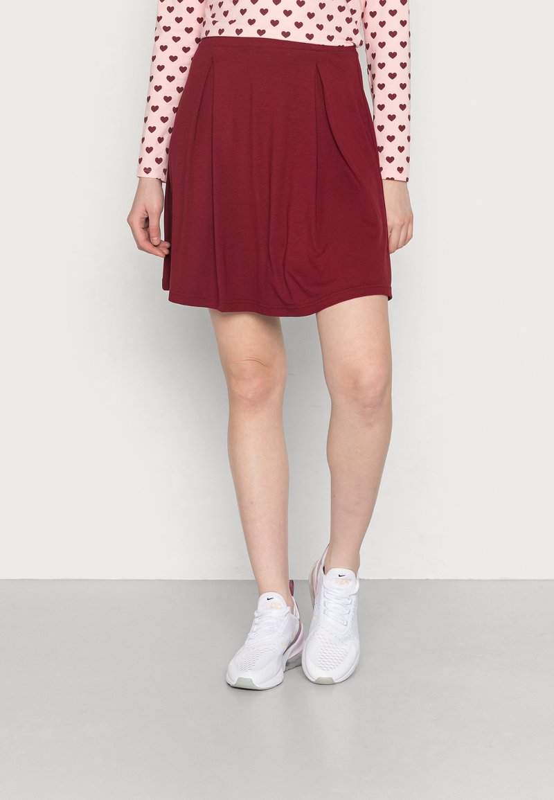 Bordeauxrode skaterrok met plooien aan de taille, gemaakt van zachte stof. Gecombineerd met witte sportschoenen en een roze top met lange mouwen en hartjespatroon.