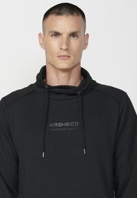 Sudadera negra con textura de espiga, cuello alto y cordón. Presenta el logo de KRSHECO en gris en el pecho. Diseño ajustado.