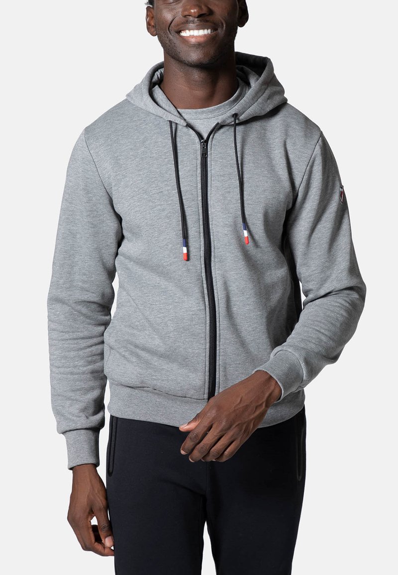 Sudadera gris con cremallera, hecha de tela suave, con capucha ajustable, mangas largas y bolsillos laterales. Detalles con puntas de cordón de colores.