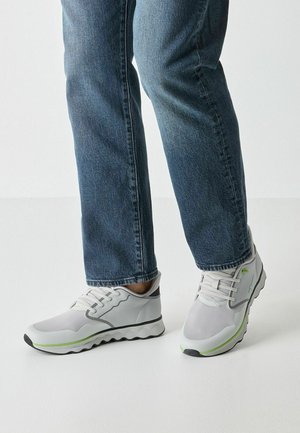 Chaussures de sport gris clair avec des panneaux texturés, des accents verts et une semelle en forme d'onde. Portées avec un jean denim bleu.