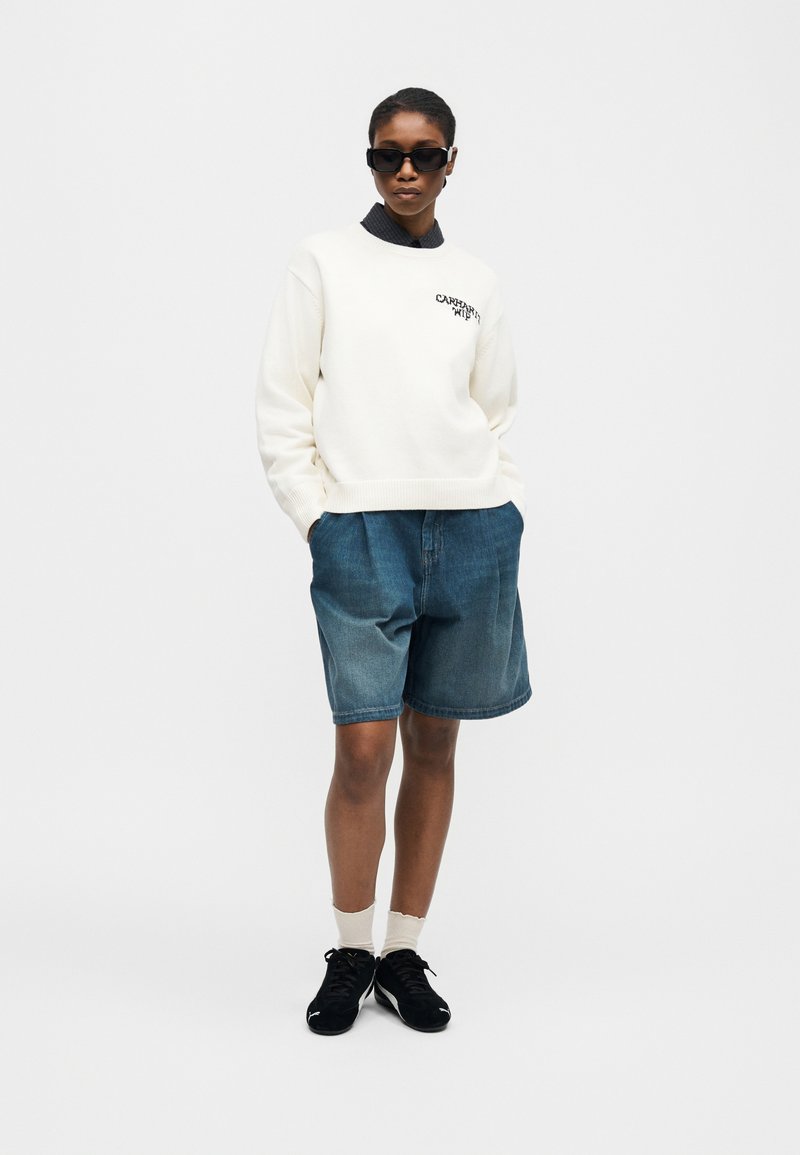 Donna con occhiali da sole neri, felpa bianca con logo, shorts di jeans blu, calzini bianchi e scarpe da ginnastica nere, in piedi con le mani in tasca.