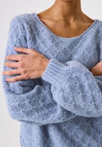 Personne croisant les bras sur la poitrine, portant un pull tricoté texturé bleu clair avec un large col.