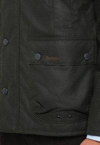 Tmavě zelená bunda s texturovaným povrchem, vybavená patentními knoflíky a náprsní kapsou vyšívanou nápisem "Barbour" hnědou barvou.