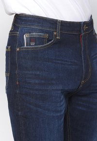 Donkerblauwe denim jeans met een recht model, zichtbare stiksels in het geel en een klein label op de achterzak.