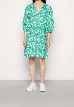 Femme portant une robe verte à fleurs longueur genou avec des manches bouffantes et des baskets blanches, debout devant un fond uni.