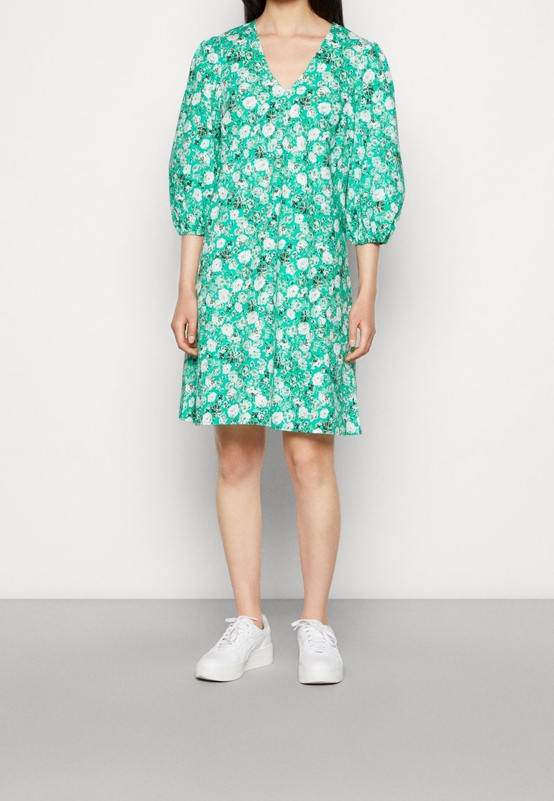 Femme portant une robe verte à fleurs longueur genou avec des manches bouffantes et des baskets blanches, debout devant un fond uni.