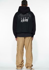 Sudadera negra extragrande con capucha, que presenta letras blancas en la parte posterior. Combinada con pantalones cargo khaki de corte holgado y zapatillas blancas.