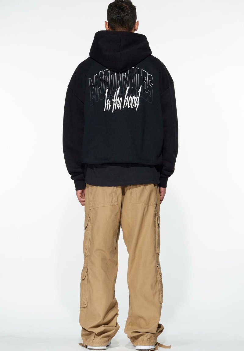 Sudadera negra extragrande con capucha, que presenta letras blancas en la parte posterior. Combinada con pantalones cargo khaki de corte holgado y zapatillas blancas.