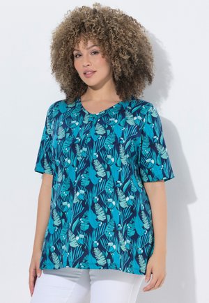 Donna con capelli ricci indossa una camicia azzurra con motivi di foglie tropicali e uccelli, abbinata a pantaloni bianchi, in piedi davanti a uno sfondo bianco.