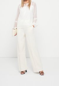 Weiße Bluse mit transparenten, gepunkteten Ärmeln, Kordelzug am Hals und elastischen Bündchen; kombiniert mit cremefarbenen, weit geschnittenen Hosen und gestreiften High Heels.