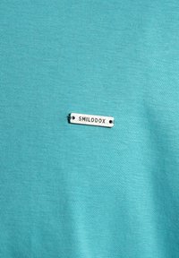 Turkos klädestyg med en liten rektangulär metalltag sydd på, med engraverat ordet "SMILODOX."