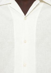Λινό πουκάμισο σε εκρού απόχρωση με διακριτική υφή, διαθέτει μυτερό γιακά και τρία ανοιχτόχρωμα κουμπιά κατά μήκος της πλακέτας.