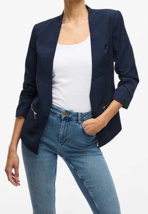 Mujer que lleva una chaqueta azul marino con bolsillos con cremallera, un top blanco sin mangas y unos vaqueros azules claros de talle alto, con una mano en el bolsillo, sobre fondo blanco.