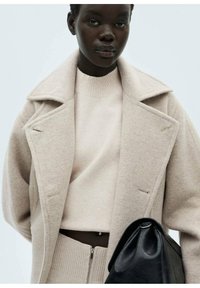 Beige Wollmischmantel mit breiten Revers, kombiniert mit einem hellbeigen Strickpullover. Schwarze Ledertasche in runder Form lehnt gegen das Outfit.