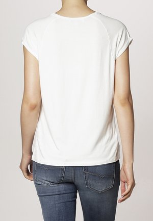 Persoon in een eenvoudig wit T-shirt met korte mouwen en blauwe jeans, bekeken vanuit de rug tegen een neutrale achtergrond.