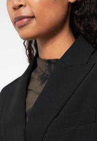 Blazer nero con rever a punta, caratterizzato da una texture liscia. Sotto, una maglia marrone con motivo, con disegni sottili visibili all'escollatura.