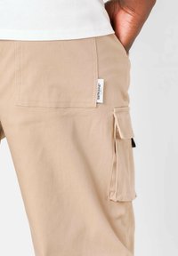 Beige cargobroek van stof met een gladde textuur, voorzien van een zijzak, patchdetails en een label dat in de tailleband is gestikt.