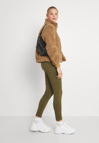 Casaco de fleece castanho com colarinho e fecho de botão, combinado com leggings verde-oliva. O modelo usa ténis brancos e uma bolsa de ombro preta.