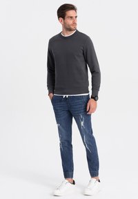 Sudadera gris con cuello redondo, combinada con vaqueros azul claro desgastados que presentan puños elásticos. Zapatillas blancas con detalles en negro completan el conjunto.