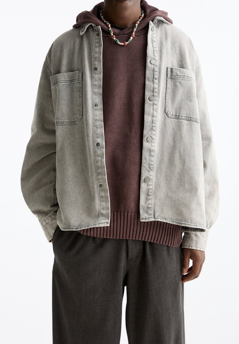 Personne portant une veste beige boutonnée sur un sweat à capuche marron et un pantalon sombre, accessoirisée d’un collier de perles.