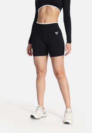 Frau trägt schwarze, hoch taillierte Sportshorts mit Logo, schwarzes langärmeliges Crop-Top mit weißem Rand und weiße Sneaker, vor weißem Hintergrund.