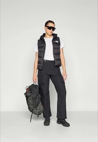 Zwarte puffer vest over een wit T-shirt, gecombineerd met zwarte cargobroek. De persoon houdt een grijze rugzak met rode accenten vast. Donkere zonnebril op.