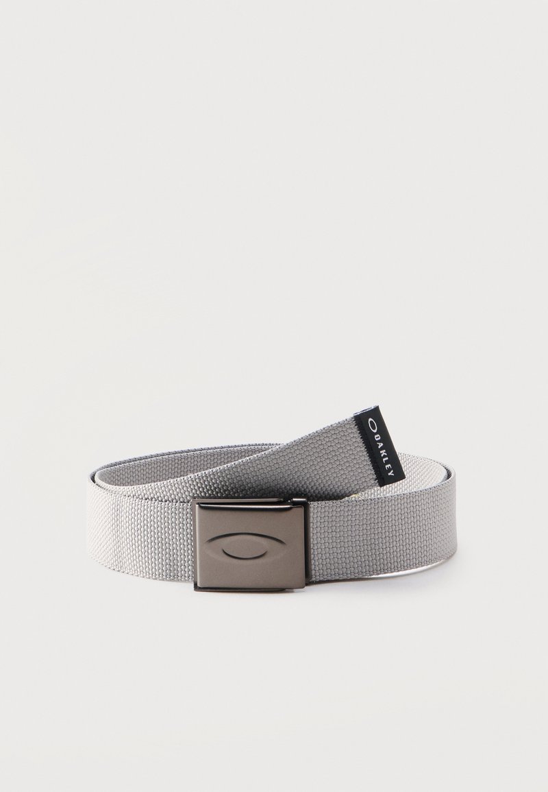 Ζώνη από υφασμάτινο ύφασμα ανοιχτού γκρι χρώματος με ματ μεταλλική αγκράφα που φέρει χαραγμένο οβάλ λογότυπο και μαύρη ετικέτα της μάρκας Oakley.