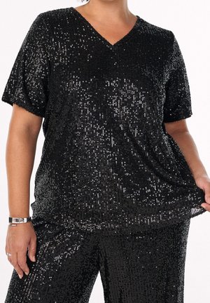 Donna che indossa un top nero a maniche corte e pantaloni ricoperti di paillettes nere lucide, tenendo l'orlo della maglia con una mano.