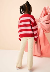 Rood en roze gestreepte gebreide sweater met een losse pasvorm, gecombineerd met crèmekleurige flared pantalon. De achtergrond toont gedrapeerd roze stof.