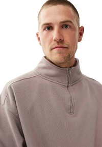 Vaaleanlaventelin värinen neljännekselle avoin vetoketjupaita, joka on valmistettu pehmeästä kankaasta. Siinä on korkea kaulus ja metallivetoketju, jossa on pieni vetotappi.