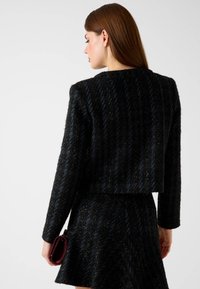 Donna con capelli castani lisci indossa un blazer nero dalla trama strutturata e una gonna coordinata, tiene una pochette bordeaux e si mostra di spalle su uno sfondo bianco.