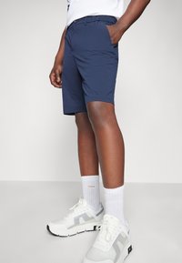 Shorts navy en tissu léger, avec une taille élastique et des poches latérales, accompagnés de chaussettes de sport blanches et de baskets gris-blanc.