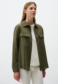 Olijfgroene overhemdjacket met een kraag, twee voorzakken, knoopsluiting en lange mouwen, gedragen over een wit topje en lichte broeken.