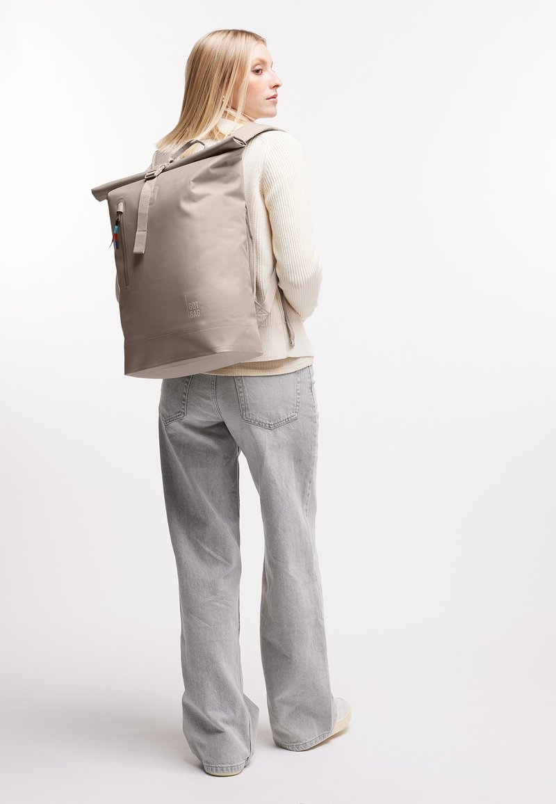 Frau mit langen blonden Haaren, die einen cremefarbenen Pullover, graue weit geschnittene Jeans und beige Sneaker trägt, und einen großen beigen Rolltop-Rucksack bei sich hat.