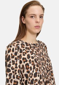 Blusa con stampa leopardata caratterizzata da un design plissé, tessuto morbido, scollatura rotonda e una palette di colori che include beige, marrone e accenti neri.