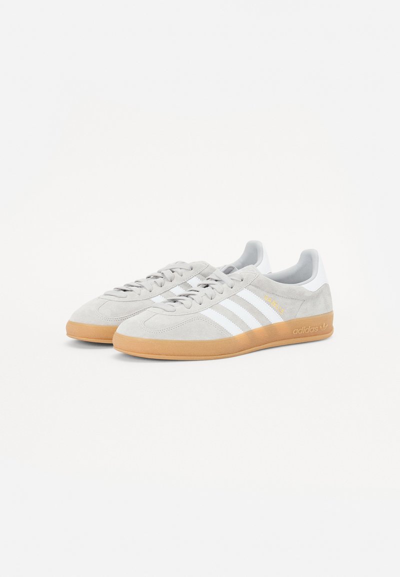 Zapatillas de ante gris con franjas blancas, suela de goma, punta redondeada y diseño de cordones. Presenta detalles dorados en el lateral.