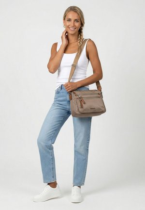 Lächelnde Frau mit geflochtenem Haar trägt ein weißes Tanktop, hellblaue Jeans, weiße Sneakers und trägt eine beige Umhängetasche vor einem einfarbigen Hintergrund.