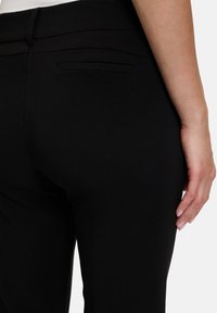 Schwarze Hosen aus einem glatten, strukturierten Stoff. Sie verfügen über eine dezente Gesäßtasche und ein tailliertes Design, das eine klare, maßgeschneiderte Silhouette präsentiert.