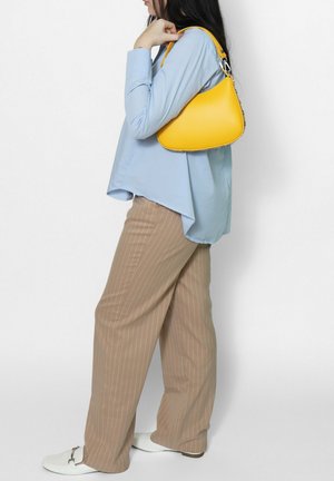 Donna con camicetta azzurra chiara e pantaloni beige a righe sottili che porta una borsa gialla sulla spalla su sfondo bianco.