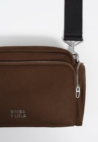 Ruda audinio petinė kuprinė su užtrauktuku, sidabriniais elementais ir nuimamu diržu su tekstūruotu juodu paviršiumi. Matomas "BIMBA Y LOLA" logotipas.