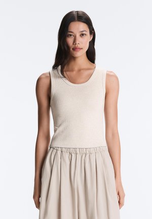 Femme aux longs cheveux foncés portant un haut beige sans manches côtelé et un pantalon beige ample, debout devant un fond clair uni.