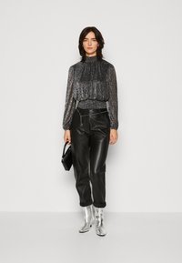 Blouse noire scintillante à manches longues et col haut, associée à un pantalon en cuir noir ajusté. Des bottines argentées et un petit sac noir complètent le look.