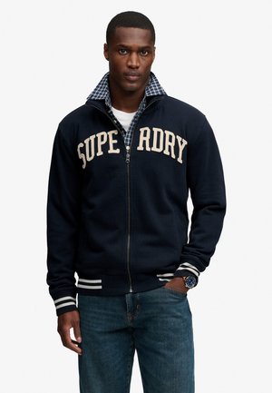 Mężczyzna w granatowej kurtce zapinanej na zamek z napisem „SUPERDRY”, pod spodem w kratę koszula, niebieskie dżinsy i zegarek na rękę, stoi z jedną ręką w kieszeni.