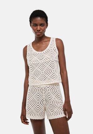 Femme portant un haut sans manches blanc en crochet et un short assorti, debout devant un fond clair uni.