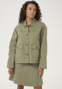 Alkūninė žalia medvilninė striukė su sagų uždarymu, dviem priekinėmis kišenėmis ir dekoratyviniais nagais ant apykaklės, derinama su atitinkamomis sijoninėmis.