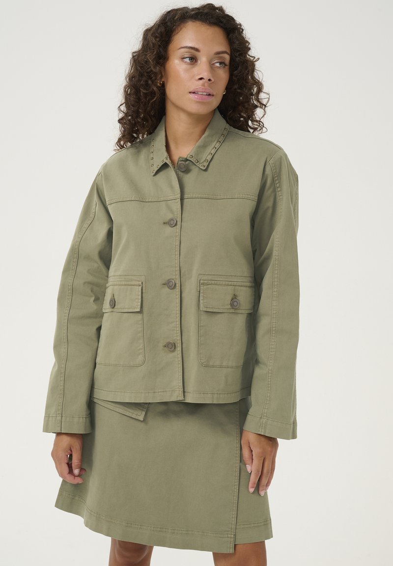 Alkūninė žalia medvilninė striukė su sagų uždarymu, dviem priekinėmis kišenėmis ir dekoratyviniais nagais ant apykaklės, derinama su atitinkamomis sijoninėmis.