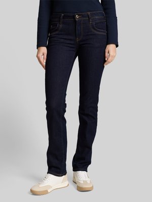 Kvinde iført mørkeblå smalle jeans og hvide sneakers, der står foran en ensfarvet grå baggrund.