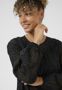 Kvinde med krøllet hår iført en sort gennemsigtig bluse med blomsterbeklædning, smilende og kigger til siden, hånd nær hendes ansigt.