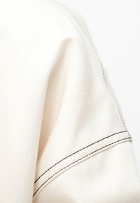 Μπεζ ύφασμα με  υφή που αναδεικνύει μια ορατή ραφή τονισμένη με σκούρα ραφές, υπογραμμίζοντας το δομημένο του σχέδιο.